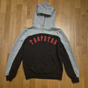 Trapstar hoodie i svart och grå - Säljer en snygg hoodie från Trapstar i svart och ljusgrått med röd logga framtill. Tröjan har huva, känguruficka och långa ärmar. Perfekt för dig som gillar streetwear och vill sticka ut med cool design.