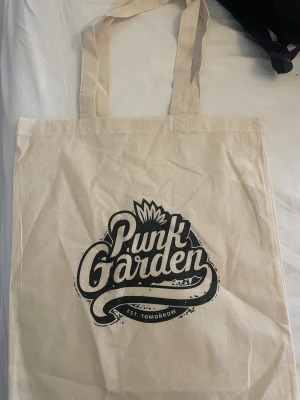Beige tygkasse Punk Garden - Snygg beige tygkasse med svart tryck från Punk Garden. Väskan har långa handtag och coolt grafiskt motiv med texten 'Punk Garden' och 'Est. Tomorrow' på framsidan. Perfekt för att bära böcker, laptop eller shopping. Enkel och stilren design.