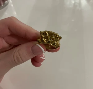 Guldfärgad ring med nitar från EDBLAD - Cool och chunky guldfärgad ring från EDBLAD med flera konformade nitar på ovansidan. Ringen har en bred design och sticker verkligen ut med sin edgy stil. Perfekt för dig som gillar statement-smycken och vill ha något som syns.