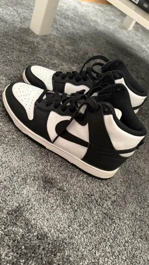 Nike Dunk High svart/vit sneakers - Säljer ett par Nike Dunk High sneakers i svart och vitt skinn med klassisk swoosh på sidorna. Skorna har svarta snören, perforerad tåbox och en vit mellansula. Perfekt för dig som gillar streetstyle och vill ha en ikonisk look.