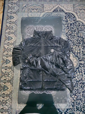 Svart pufferjacka från Trapstar - Säljer en svart pufferjacka från Trapstar med storleken S. Glansig finish och hög krage. Jackan har huva, dragkedja framtill och två fickor med tryckknappar. Trapstar-logga på bröstet och broderad text på ryggen. Perfekt för kalla dagar.