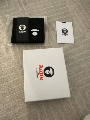 Svart plånbok från Aape by A Bathing Ape - Snygg svart plånbok från Aape by A Bathing Ape med broderad logga framtill. Plånboken har flera kortfack, myntficka med tryckknapp och är tillverkad i slitstark polyester. Kommer i originalförpackning och med tillhörande kort.