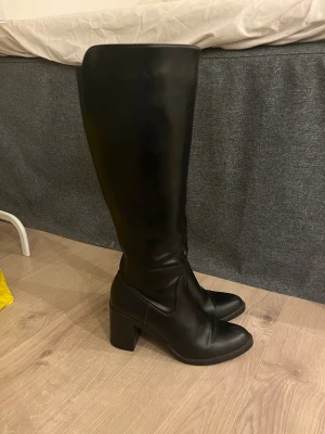 Svarta höga stövlar med klack - Snygga svarta stövlar i skinnimitation med hög klack och spetsig tå. Stövlarna går upp till knäet och har dragkedja på insidan för enkel påtagning. Perfekt till höst och vinter för en stilren look.