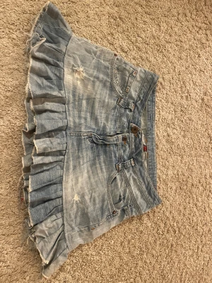 Ljusblå volang jeanskjol från Huca Denim - Supersöt ljusblå jeanskjol från Huca Denim med volangkant och slitna detaljer. Klassisk femficksmodell, bälteshällor och knappar framtill. Kjolen har en cool, lite ruffig look och är perfekt för dig som gillar denim med attityd.