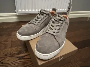 Gråa Louboutin skor - Snygga grå sneakers från Christian Louboutin i mjuk mocka med klassisk vit sula och ikonisk röd undersida. Skorna har rund tå, snörning och diskret branding. Perfekta för dig som vill ha en lyxig och stilren look med en touch av Paris.