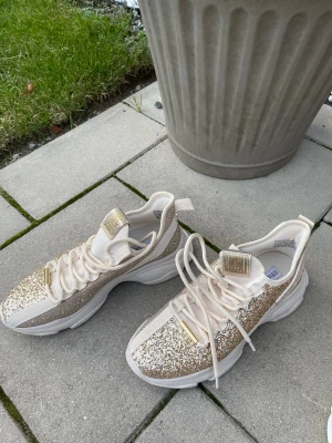 Glittriga sneakers i guld och beige - Säljer ett par Steve Madden sneakers med chunky vit sula och glittriga detaljer i guld och beige. Skorna har snörning med guldfärgade metalldetaljer och en fyrkantig glittrig patch på plösen. Perfekt för dig som gillar att sticka ut med din stil. Stl 40 små i stl / dom en 39