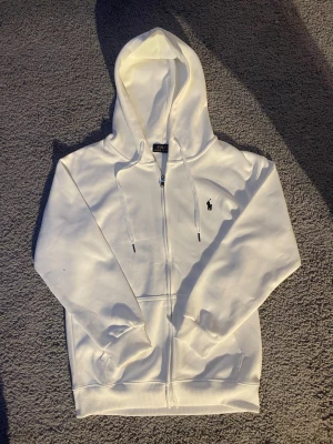 Vit hoodie från Polo Ralph Lauren  - Vit hoodie från Polo Ralph Lauren i storlek M men den sitter litte mindre. Tröjan har huva med snörning, hel dragkedja och klassisk broderad logga på bröstet. Tillverkad i mjuk bomull med ribbade muddar och fickor framtill. Perfekt för chill dagar och snygg streetstil.