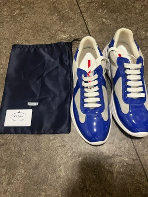 Blå vita Prada cups - Snygga sneakers från Prada i blått och vitt med röda detaljer. Skorna har en kombination av glansigt skinn och mesh, vita snören och rund tå. Klassisk chunky sula och diskret Prada-logga baktill. Perfekta för dig som vill sticka ut med stil. Storlek 45