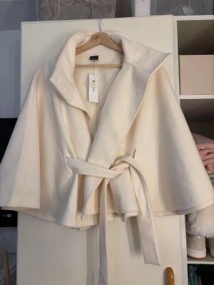 Vit capejacka med bälte från Gina Tricot - Stilren vit capejacka från Gina Tricot med bred krage och knytbälte i midjan. Jackan har en loose passform och är tillverkad i mjukt material som känns lyxigt. Perfekt för dig som vill ha en trendig och elegant look.