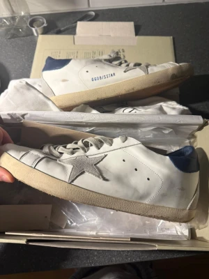 Golden Goose vita sneakers med blå detalj - Golden Goose sneakers i vitt skinn med grå mockastjärna på sidan och blå hälkappa. Skorna har grå snörning och en beige sula med vintage-look. Perfekta för dig som gillar en avslappnad men trendig stil.