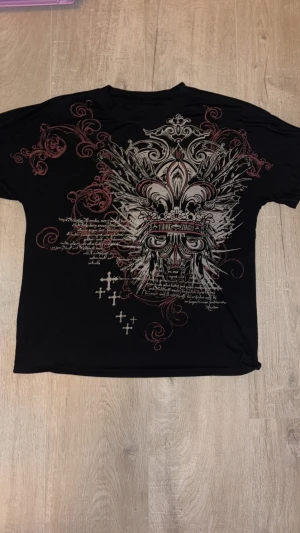 Svart t-shirt med tryck - Svart t-shirt med korta ärmar och stort barockinspirerat tryck i silver och rött framtill. Designen har detaljer som kors, snirkliga mönster och text, vilket ger en edgy och unik vibe. Perfekt för dig som gillar statement-plagg med mycket attityd.