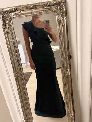 Mörkgrön långklänning med volang - Säljer en elegant mörkgrön långklänning med oneshoulder och volangdetaljer upptill. Klänningen har en figurnära passform och är perfekt för bal eller fest. Aldrig använd med prislappen kvar. 