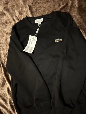 Svart sweatshirt från Lacoste M - Svart sweatshirt från Lacoste i regular fit med klassisk rund hals och det ikoniska gröna krokodilmärket broderat på bröstet. Tröjan har ribbade muddar vid ärmslut och nederkant. Perfekt för en chill och stilren look.
