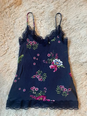 Mörkblå blommig Tezenis  linnetopp med spets - Supersöt mörkblå linnetopp med tunna axelband och blommigt mönster i rosa, grönt och vitt från Tezenis.Toppen har fina spetsdetaljer längs halsringningen och nederkanten som ger en romantisk vibe. Perfekt att styla med jeans eller kjol för en fräsch look.