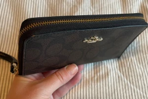 Svart plånbok med handledsrem från Coach - Snygg svart plånbok från Coach med diskret mönster och guldfärgad dragkedja. Plånboken har en praktisk handledsrem och flera fack för kort och kontanter. Materialet är skinn och insidan är stilren med plats för allt du behöver.