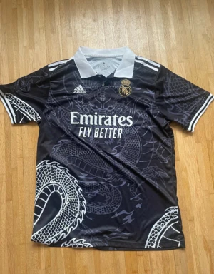 Svart Real Madrid fotbollströja Adidas - Snygg svart Real Madrid fotbollströja från Adidas med vit krage och detaljer. Tröjan har ett stort drakmönster över hela framsidan och baksidan, samt klubbmärke och sponsortryck på bröstet. Kortärmad och tillverkad i lätt, ventilerande material.