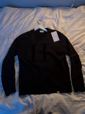 Svart sweatshirt från Les Deux - Svart sweatshirt från Les Deux med stort, stilrent II-tryck på bröstet. Tröjan har rund hals, långa ärmar och ribbade muddar vid ärmslut och nederkant. Materialet är mjuk bomull som känns skön mot huden. Perfekt för en clean och modern look. Är en S men passar en person med M