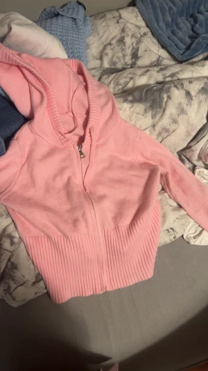 Rosa stickad hoodie med dragkedja - Säljer en ljusrosa stickad hoodie med dragkedja framtill och ribbade muddar vid ärmslut och nederkant. Tröjan har huva och är långärmad, perfekt för dig som gillar mysiga och trendiga plagg. Passar dig som vill ha en chill och söt look.