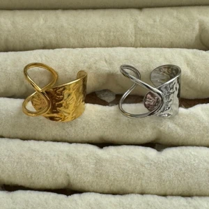 Två breda ringar i guld och silver - Säljer två snygga breda ringar med hamrad yta, en i guld och en i silver. Båda har en cool öppen design med en snirklig detalj framtill som ger dem en unik look. Perfekt för dig som gillar statement-smycken och vill sticka ut.