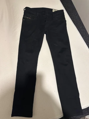 Svarta Diesel D-Bazer tapered jeans - Säljer ett par svarta Diesel D-Bazer jeans med tapered passform. Snygga detaljer som metallmärke och Diesel Industry-etikett vid fickan. Jeansen har klassisk femficksdesign och knäppning framtill. Stretchigt material för extra komfort.