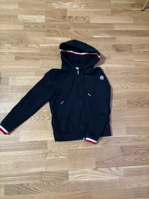 Svart hoodie från Moncler med dragkedja - Svart hoodie från Moncler med hel dragkedja, huva och snörning. Röd och vit randig detalj på huva och ärmslut samt Moncler-logga på ärmen. Perfekt för en stilren och sportig look.