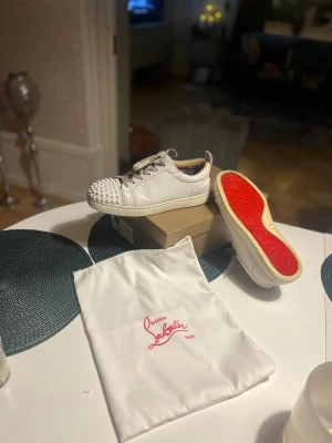 Christian Louboutin vita sneakers med nitar - Säljer ett par vita sneakers från Christian Louboutin med ikonisk röd sula och coola nitar på tån. Skorna har snörning och är tillverkade i skinn, vilket ger en lyxig känsla. Perfekt för dig som vill sticka ut med en edgy och exklusiv stil. Äkta naturligtvis