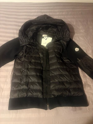 Svart Moncler dunjacka med huva - Svart dunjacka från Moncler med quiltad front, stickade ärmar och ribbade muddar. Jackan har huva, dragkedja framtill och Moncler-logga på ärmen. Perfekt för kyliga dagar och riktigt snygg streetstil.