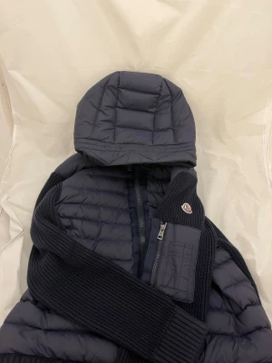 Mörkblå Moncler pufferjacka med huva - Snygg mörkblå pufferjacka från Moncler med stickade ärmar och detaljer. Jackan har en vadderad huva, dragkedja framtill och en bröstficka med dragkedja. Perfekt för kyliga dagar och riktigt stilren med Moncler-logga på ärmen.
