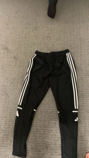 Svarta Adidas träningsbyxor M - Svarta träningsbyxor från Adidas i storlek M med vita ränder längs sidorna och logga på benen. Byxorna har elastisk midja och är gjorda i polyester för skön passform. Perfekta för sport eller chill, med smal passning och snygga detaljer.