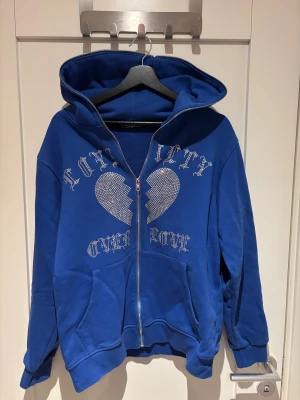 Blå hoodie med strass från Trotherz - Blå hoodie från Trotherz med hel dragkedja, även över huvan. Framsidan har ett stort strassmönster med text och ett brustet hjärta. Tröjan är i mycket bra skick, ny skick i stort sätt. 