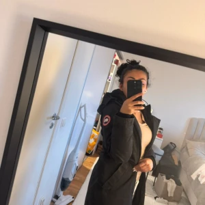 Svart parkajacka från Canada Goose - Svart parkajacka från Canada Goose med klassisk rödvit logga på ärmen. Jackan har huva med snörning, långa ärmar och praktiska fickor framtill. Snygg, stilren och perfekt för kalla dagar. Passformen är normal och jackan är tillverkad i slitstark polyester.- Päls finns med !