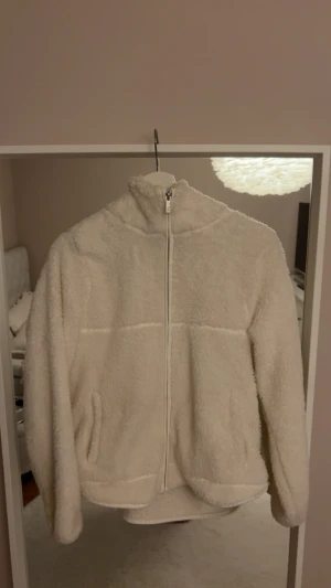 Vit teddyjacka med dragkedja från lager 157 - Mysig vit teddyjacka med hög krage och hel dragkedja framtill. Jackan har två fickor på sidorna och är tillverkad i fluffigt fleece-material som ger en riktigt skön och avslappnad vibe. Perfekt för kyliga dagar när du vill vara både varm och stylish.