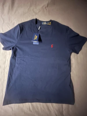 Mörkblå t-shirt från Polo Ralph Lauren - Snygg mörkblå t-shirt från Polo Ralph Lauren i mjuk bomull. Klassisk passform med rund hals och korta ärmar. På bröstet finns det ikoniska röda Polo-logot broderad. Perfekt för en clean och stilren look.