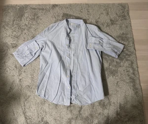 Ljusblå henley skjorta från Zara - Snygg ljusblå henley skjorta från Zara med klassisk ståkrage och knappar framtill. Skjortan har uppvikbara ärmar med knappdetaljer och är tillverkad i mjuk bomull som känns skön mot huden. Perfekt för dig som gillar stilrena och enkla plagg.