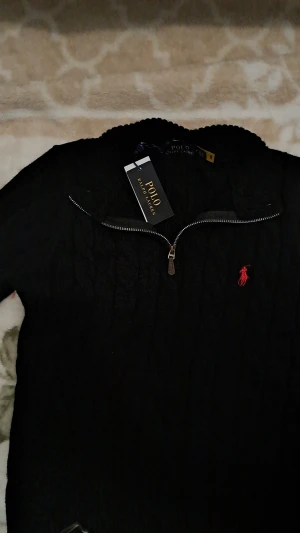 Svart stickad tröja Polo Ralph Lauren - Svart stickad tröja från Polo Ralph Lauren med kabelstickat mönster och röd broderad logga på bröstet. Tröjan har lång ärm, ribbade muddar och en snygg half zip-dragkedja vid halsen. Perfekt för dig som gillar klassisk stil med en modern touch. Helt ny med etikett på