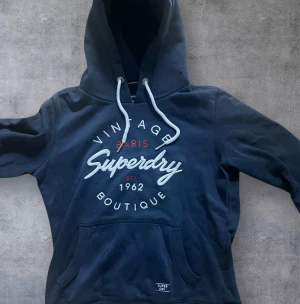 Mörkblå Superdry hoodie med tryck - Snygg mörkblå hoodie från Superdry med stort broderat och tryckt vintage-inspirerat motiv på bröstet. Tröjan har huva med vita snören, känguruficka och ribbade muddar. Perfekt för dig som gillar streetwear och vill ha en chill look.