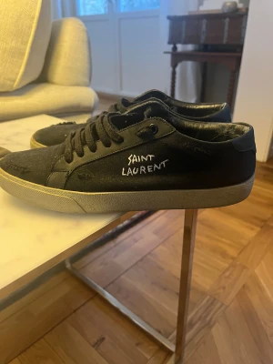 Svarta Saint Laurent sneakers - Svarta sneakers från Saint Laurent med broderad logga på sidan. Skorna har snörning, rund tå och platt sula. Ovandelen är i textil med detaljer i skinn, insidan är fodrad med svart skinn. Snygg och stilren design med kontrasterande vit sula. Däremot har skorna sett bättre dagar än nu så dessa skor passar bra till den som vill fräscha upp dem. Box medkommer.