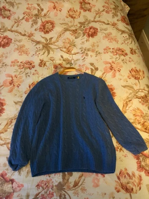 Blå kabelstickad tröja Polo Ralph Lauren - Snygg blå kabelstickad tröja från Polo Ralph Lauren med rund hals och diskret logga på bröstet. Tröjan har långärmade ribbade muddar och är gjord i mjuk merinoull. Perfekt för dig som gillar klassisk stil med en modern touch.