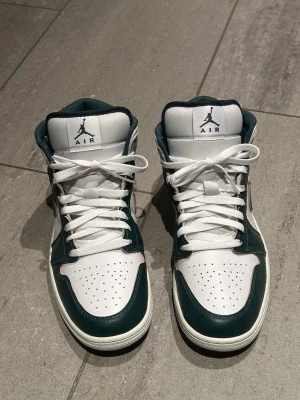 NYA - Nike Air Jordan 1 Mid vit/grön - Nike Air Jordan 1 Mid sneakers i vitt och mörkgrönt skinn med klassisk snörning och perforerad tå. Swoosh-logga på sidan och Air Jordan-logga på plösen. Skorna har en rund tå och platt sula, perfekt för dig som gillar streetwear och snygg design.