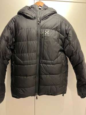 Svart dunjacka Haglöfs Belay II M - Svart dunjacka från Haglöfs, modell Belay II. Jackan har huva med snörning, flera dragkedjeförsedda fickor och tydlig logga på bröstet. Puffig design med quiltade paneler och tvåvägsdragkedja framtill. Perfekt för kalla dagar.