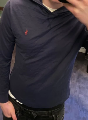 Marinblå hoodie från Polo Ralph Lauren - Snygg marinblå hoodie från Polo Ralph Lauren med klassisk röd broderad logga på bröstet. Tröjan har huva, långärmad design och är gjord i mjuk bomull. Perfekt för alla årstider! Storleken är XS