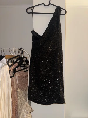 Svart paljettklänning oneshoulder - Supersnygg svart kort paljettklänning med oneshoulder-design. Klänningen är täckt av glittrande paljetter. 