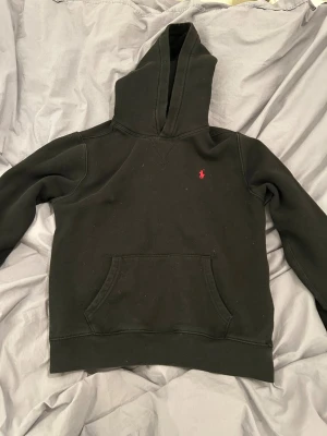 Svart hoodie från Polo Ralph Lauren - Svart hoodie från Polo Ralph Lauren med klassisk röd logobrodyr på bröstet. Tröjan har huva, magficka och ribbade muddar vid ärmslut och nederkant. Perfekt för chill dagar och enkel att matcha med jeans. Ny pris 1300. Strl L (14-16) OBS skriv till mig innan köp.