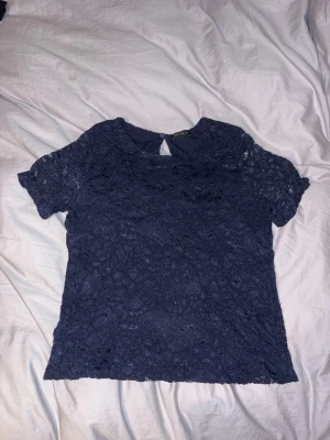 Mörkblå spetstopp - Säljer en mörkblå topp med korta ärmar och vacker spets över hela toppen. Toppen har en rund halsringning och en liten knapp i nacken med öppning. Perfekt för dig som gillar detaljer och stilren look.