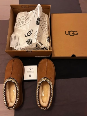 UGG tasman - Säljer ett par bruna slip-on tofflor från UGG med mjuk fårullsfoder och ovandel i mocka. Skorna har en dekorativ kant med zigzag-mönster i vitt och gult, samt en platt sula som ger skön komfort. Perfekta för att hålla fötterna varma och stilrena hemma.