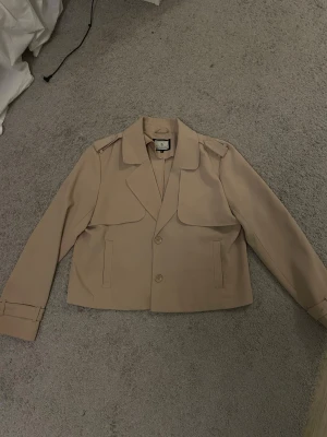 Beige kort trenchcoat från Flash Woman - Stilren beige kort trenchcoat, (kort kappa i tunt material) från Flash Woman med klassisk krage, axelklaffar och två knappar framtill. Jackan har raka ärmar med spänndetaljer vid handlederna och raka fickor. Perfekt för dig som gillar en clean och tidlös look.