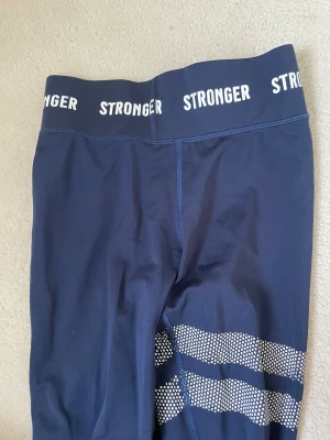Mörkblå leggings från Stronger - Snygga mörkblå leggings från Stronger med vit logga på midjan och coola prickiga detaljer på ena benet. Stretchigt material som sitter tight och är perfekt för träning. 