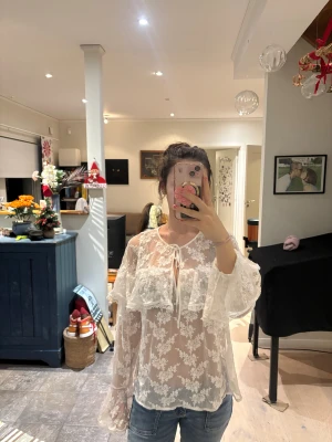 Vit blommig spetsblus med volang - Säljer en superfin vit blus i transparent spets med blommigt mönster. Blusen har långa, vida ärmar och volangdetaljer framtill samt knyt vid halsen. 