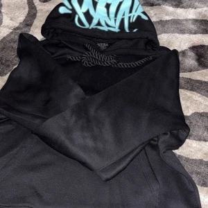 Svart hoodie SYna world med turkos print - Svart hoodie från SYNÅL i storlek M med fett turkos graffiti-print på huvan. Tröjan har snörning vid huvan och en klassisk känguruficka framtill. Materialet är mjukt och känns som bomull eller polyester, perfekt för chill dagar.