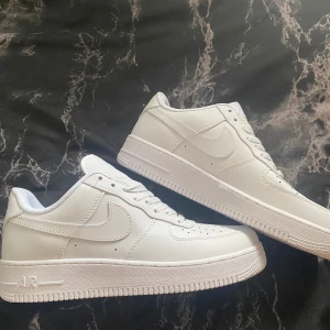 Nike Air Force 1 helvita sneakers - Klassiska Nike Air Force 1 i helvitt skinn med ikonisk swoosh på sidorna och broderad Nike Air-logga bak. Lågt skaft, perforerad tå och platt sula. Perfekta för en clean och stilren look. Skosnören i vitt och diskreta detaljer för en tidlös vibe. Boxen är vikt. 